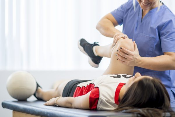 A female high school volleyball player is sitting at the doctors office to get a look at her injury. Her physical therapist is working on her knee.